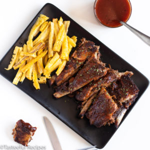 Overhead shot of baked bbq ribs and plantain fries on a platter with bbq sauce on the side and bbq ribs on one side