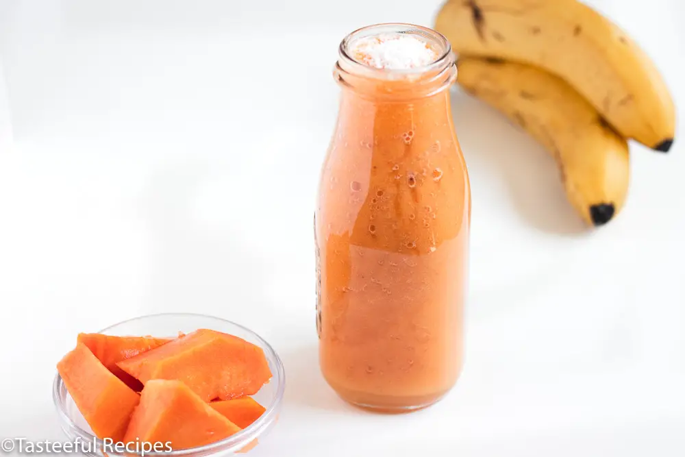 Straight on shot of a dairy free papaya smoothie in a glass bottle with bananas and sliced papaya in the background