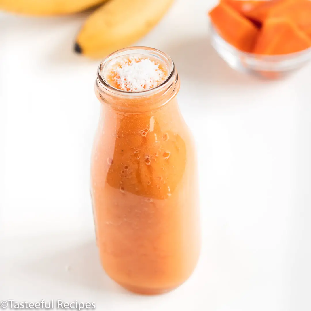 Angled shot of a dairy free papaya smoothie in a glass bottle with bananas and sliced papaya in the background